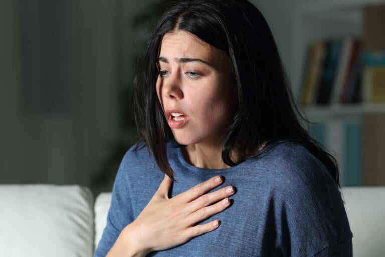 Consulta realizada correctamente La imagen muestra el primer plano de una mujer joven con cabello oscuro, su expresión facial denota angustia o dificultad para respirar. Tiene la boca ligeramente abierta, los ojos muy abiertos y las cejas fruncidas, y una de sus manos está apoyada en su pecho. La iluminación es tenue, lo que sugiere un ambiente oscuro o nocturno, y el fondo está borroso, pero se intuye que está en un interior, posiblemente en un sofá o sillón. La escena transmite una sensación de preocupación, pánico o malestar físico.